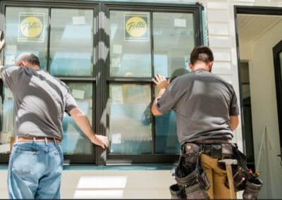 Two workers installing large black windows in a home exterior.