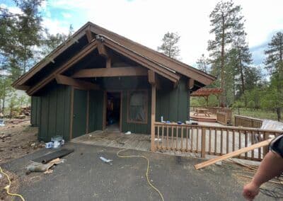 Green rustic cabin with wooden beams and a deck surrounded by trees in a natural setting.