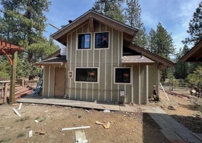 Modern two-story house with green exterior, large windows, and surrounded by pine trees.