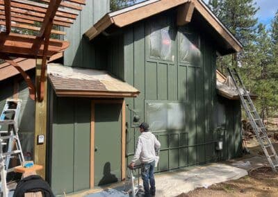 Person painting a green modern house exterior with ladders and tools nearby in a forested area.