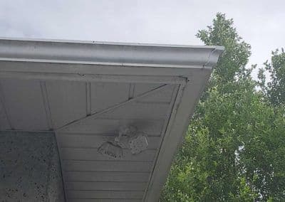 Ceiling fan mounted under a house eave with greenery in the background.