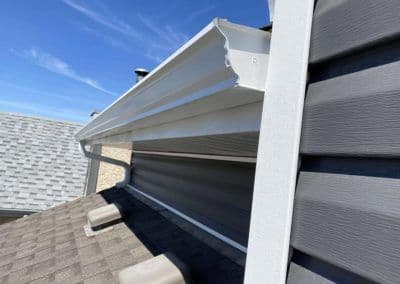 White metal gutter installed on a roof with gray shingles and clear blue sky backdrop.