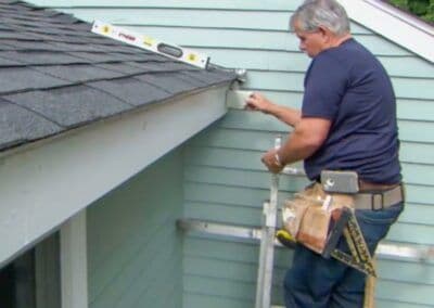 Man installing a gutter on a house, using a ladder and tools for home maintenance.