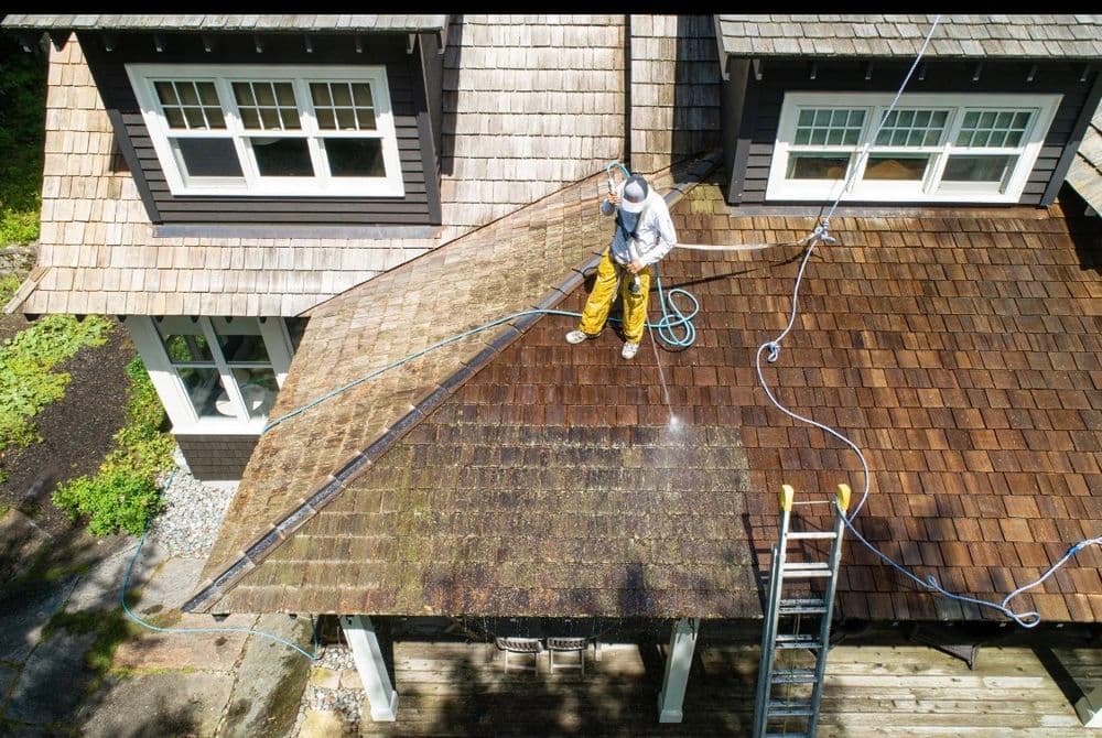 Man power washing a wooden roof, revealing clean shingles on a sunny day.